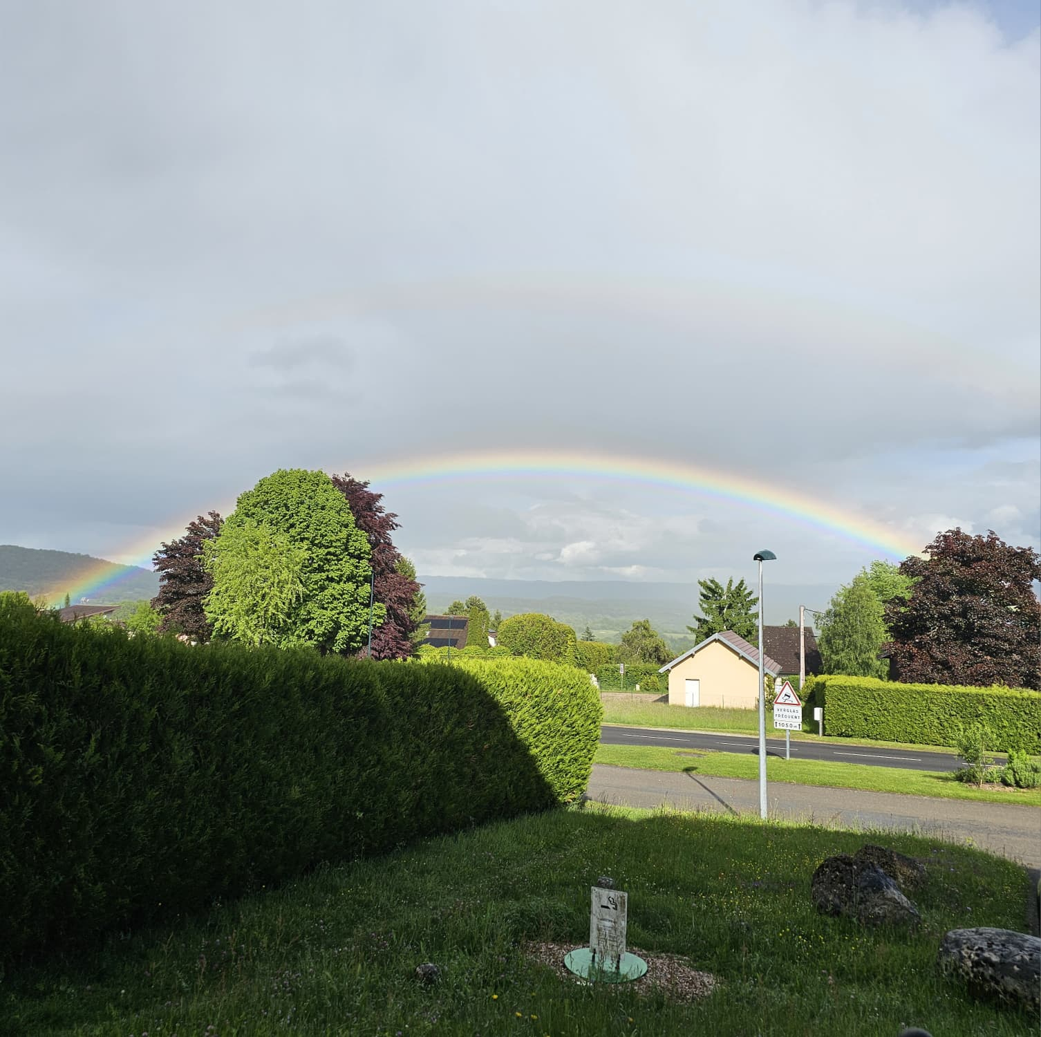 un arc en ciel au dessus d'un quartier résidentiel avec quelques pavillons et pas mal de verdure, sur un arrière-plan de basse montagne